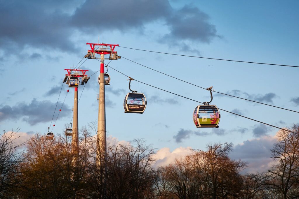 Seilbahn mit vier zu sehenden Gondeln. Wolken am Himmel und Bäume am unteren Bildrand.