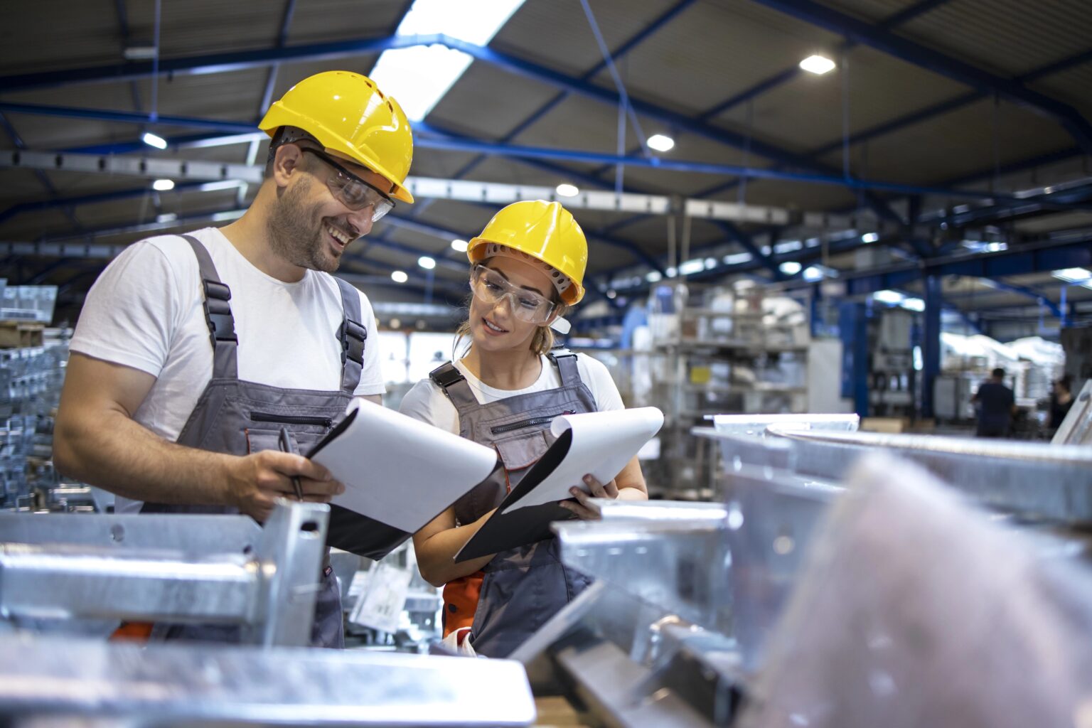 Mann und Frau in Arbeitskleidung und Helm blicken auf Unterlagen auf ihren Klemmbrettern